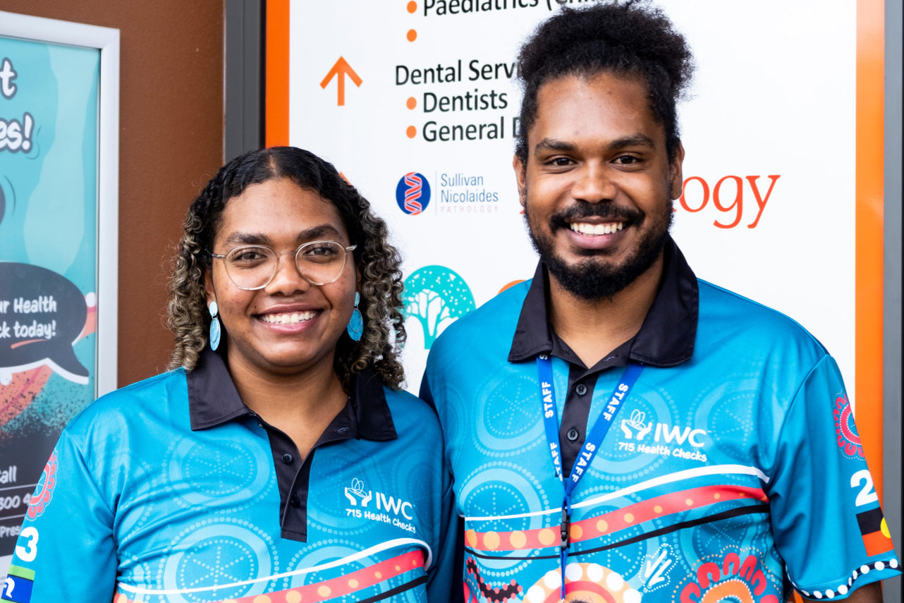 An Indigenous man models a 715 Health Check Shirt while facing the camera, standing next to an indigenous woman modelling the same shirt, facing away from the camera
