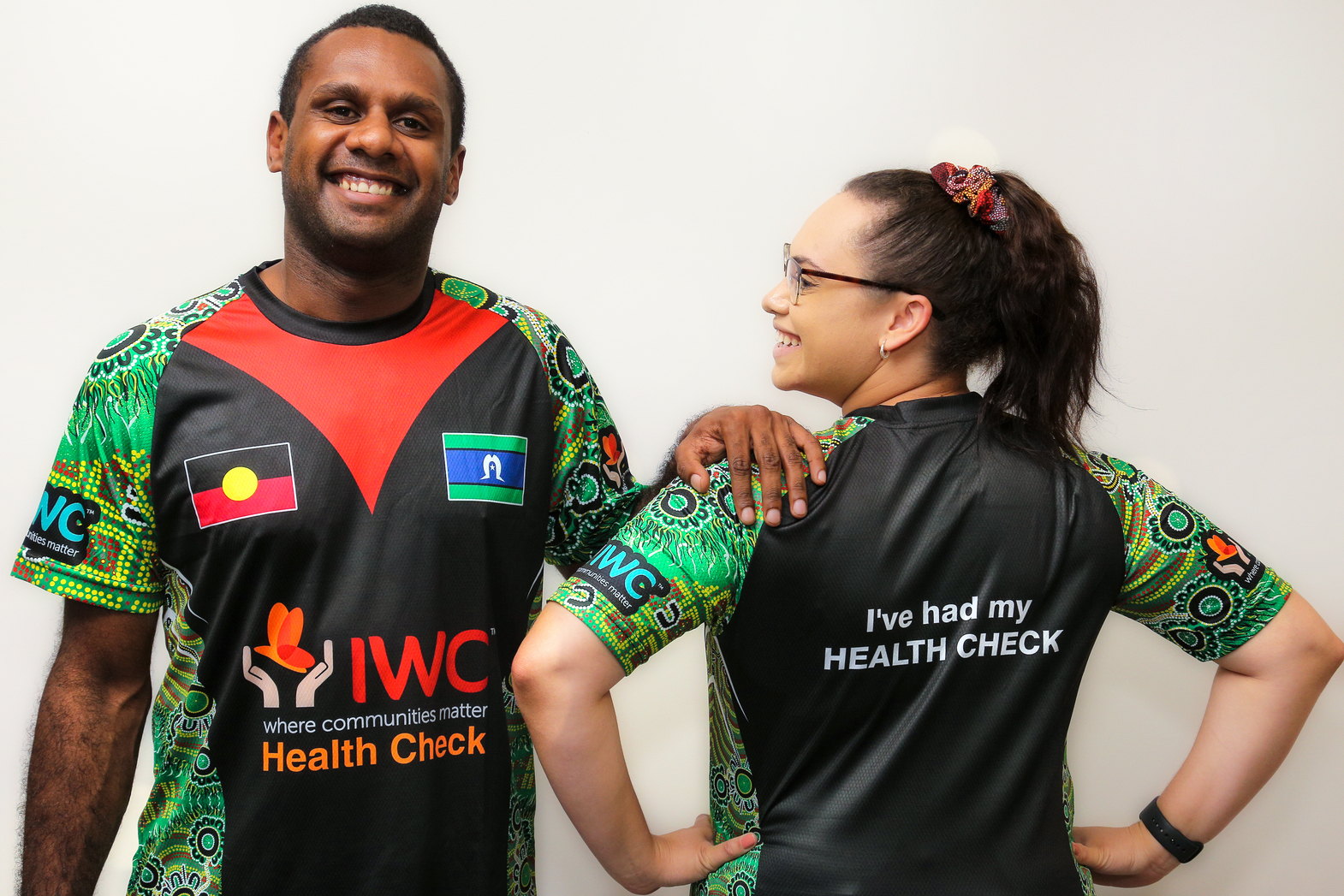 An Indigenous man models a 715 Health Check Shirt while facing the camera, standing next to an indigenous woman modelling the same shirt, facing away from the camera