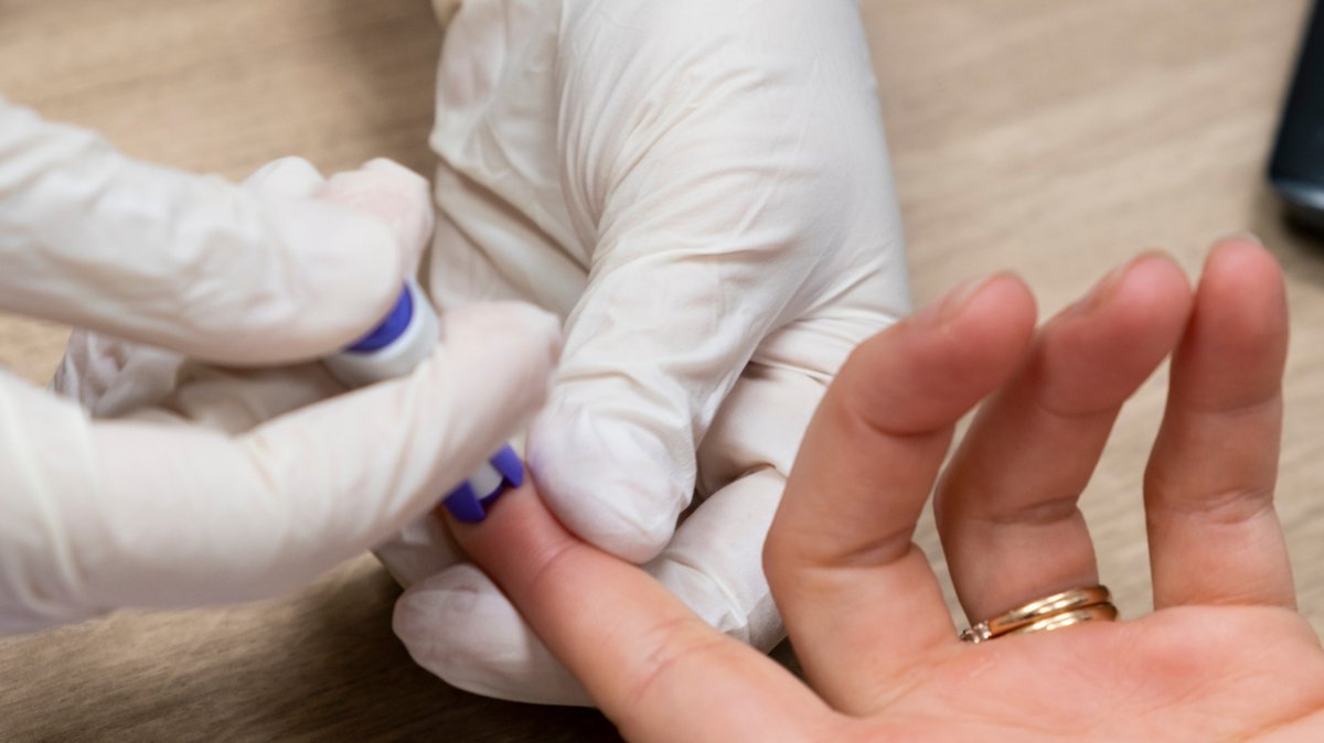 Gloved hand pricking the finger of a patients hand for a blood sugar reading.