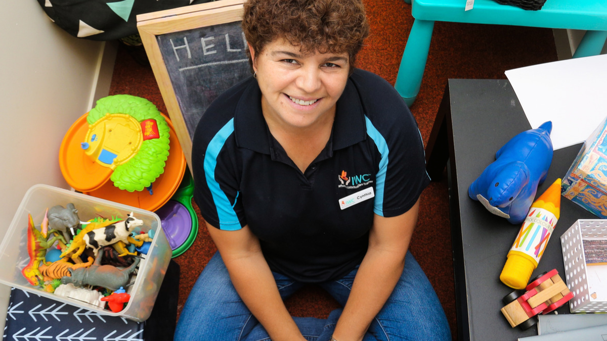 Cynthia George sits on floor crossed legged surrounded by toys smiling up at camera