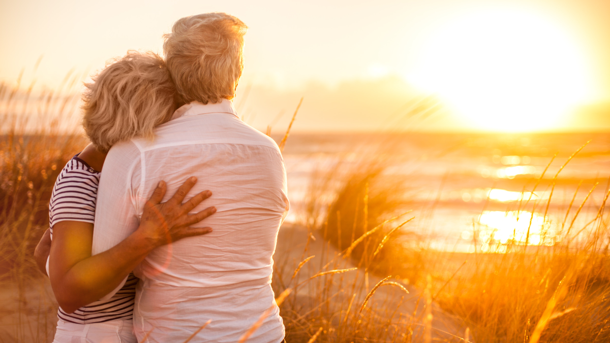 An older couple stands looking at a golden sunset over the beach while embracing