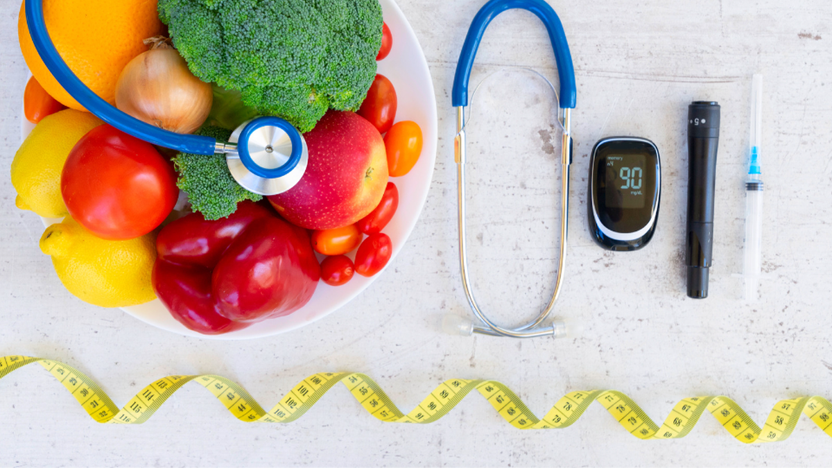 Bowl of fruit and vegetables beside stethoscope, blood glucose montior, insulin injector and a measuring tape below