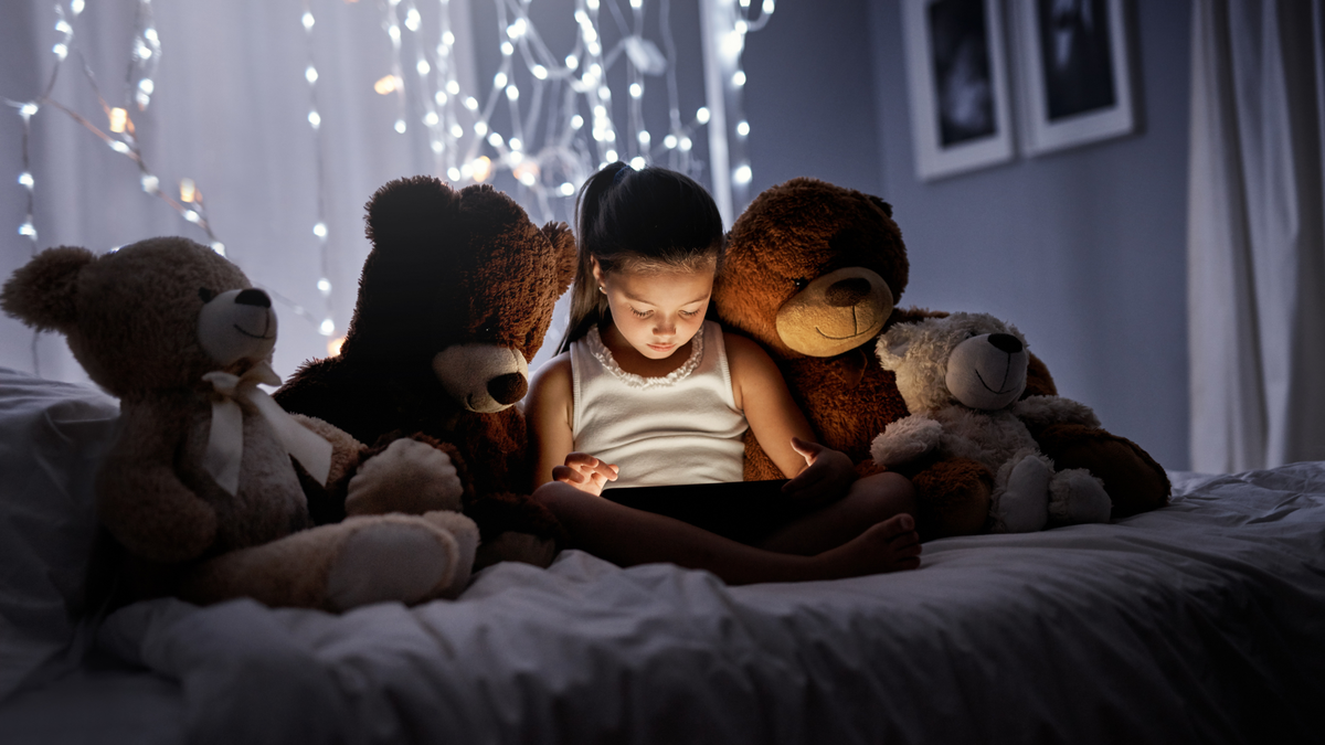 young girl watches screen on bed with large teddy bears.